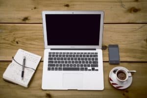 Laptop with a blank screen, a smartphone, a notepad with a pen, and a cup of coffee on a wooden desk.