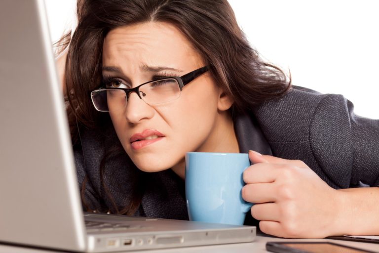 A woman with glasses holding a cup looks closely on a laptop with a frowning face