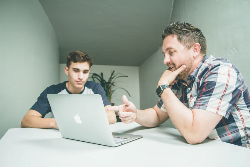 Two men sitting, looking at an opened Macbook placed on a table