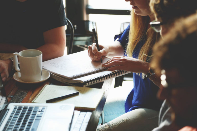 Around four people sitting around a table with a cup, two notebooks, and a laptop place on it, one of them is writing on one notebook