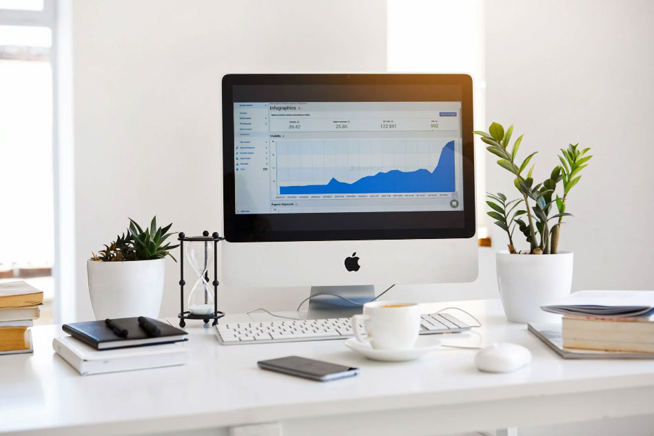 A table with iMac desktop, small plants, books, phone, and a cup on top