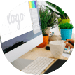 A desktop setup with an open laptop displaying a logo design, surrounded by potted plants and a person's hand holding a coffee cup.