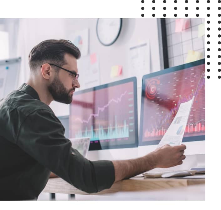 Man with beard analyzing financial documents in front of multiple computer screens displaying graphs.