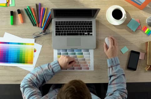 Person working at a desk with a laptop, mouse, color swatches, colored pens, highlighters, coffee mug, smartphone, and sticky notes laid out.