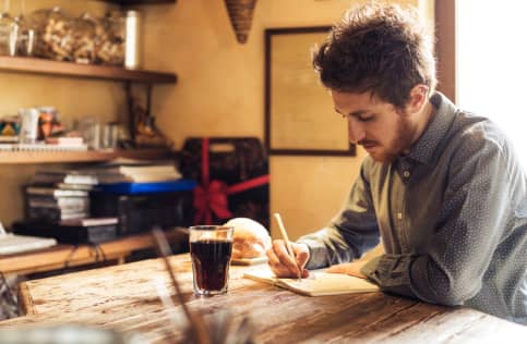 A person with a beard and casually dressed is sitting at a rustic wooden table, writing in a notebook with a drink and bread roll nearby.